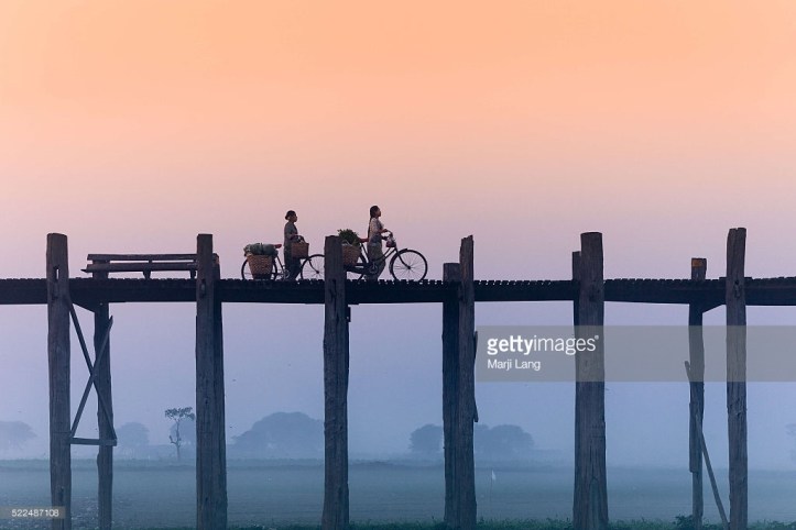 AMARAPURA, MANDALAY, MYANMAR, AMARAPURA, MANDALAY, MYANMAR - 2013/12/28: Cycling women on the U Bein bridge in Amarapura by a foggy and misty morning, near Mandalay, Burma (Myanmar). (Photo by Marji Lang/LightRocket via Getty Images)